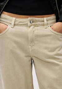 Άτομο που φοράει μπεζ τζιν με τα χέρια στις τσέπες και ένα μαύρο κοντό τοπ, που δείχνει την κοιλιά και τη λεπτομέρεια με φερμουάρ στο μπουφάν.