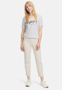 Lichtgrijze T-shirt met korte mouwen met "HAPPY" gedrukt in zwart en groen. Gecombineerd met beige slim-fit broek en witte sneakers.