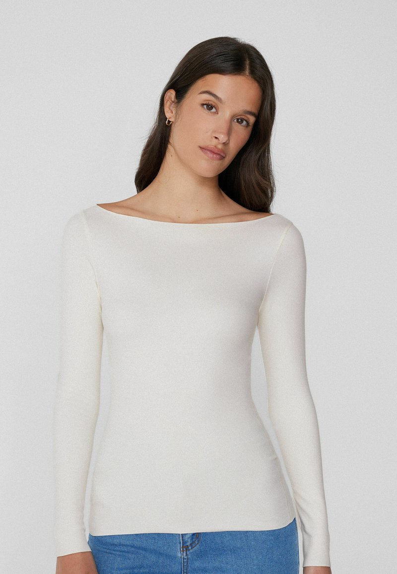 Langwerpige, off-white top met een bootnek ontwerp, gemaakt van een gladde stof, fitted pasvorm, gecombineerd met blauwe spijkerbroeken.