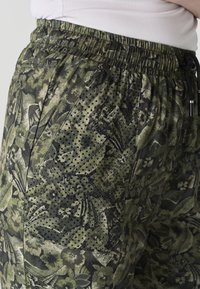 Gros plan sur un pantalon à motifs floraux verts et noirs avec une ceinture élastique et des poches latérales, porté avec un haut blanc.