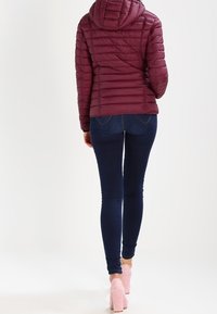 Burgundowa kurtka puchowa z kapturem o pikowanym wzorze, w połączeniu z ciemnoniebieskimi dżinsami skinny i jasnoróżowymi szpilkami.