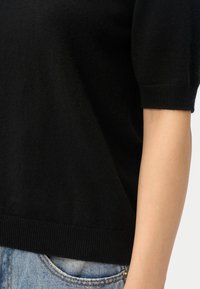 Svart kortärmad stickad tröja kombinerad med ljusblå jeans, visar en närbild av bålen och höger arm.