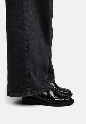 Mocasines de charol negro usados con pantalones negros de pierna ancha, mostrados de lado sobre un fondo liso.