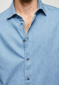 Chemise en denim bleu clair avec un col boutonné, des boutons sur le devant et une texture douce. Détail notable : un plastron cousu et une coupe décontractée.