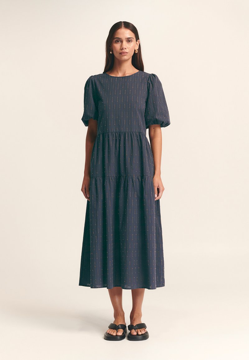 Abito midi navy con maniche a palloncino, design a strati, motivo sottile e texture morbida. Indossato con sandali neri con cinturini larghi.