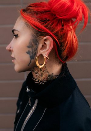 Person with bright red hair in a bun, floral face and neck tattoos, large gold geometric hoop earrings, and a black jacket with a zipper.