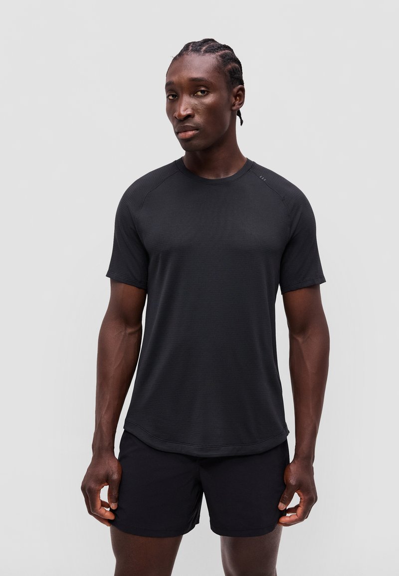 Mann mit geflochtenem Haar, der ein schlichtes schwarzes kurzärmeliges Shirt und schwarze Shorts trägt, steht vor einem neutralen grauen Hintergrund.