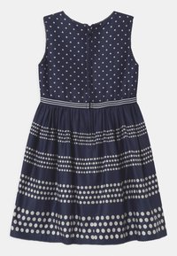 Laivastonsininen ilman hiuksia oleva mekko, jossa on valkoisia pilkkuja ja vaaka-raitakuosia. Mekossa on pyöreä kaula-aukko ja takana vetoketjukiinnitys.