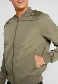 Uomo con una giacca bomber verde oliva, con zip frontale e mano nella tasca destra, in piedi su uno sfondo chiaro e semplice.