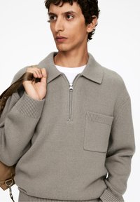 Grauer Strickpullover mit Reißverschlusskragen, gerippten Bündchen und Saum sowie einer frontalen Paspeltasche. Getragen über einem weißen T-Shirt, gehalten mit einem braunen Gürtel.