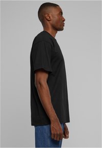 Tricou negru din bumbac, cu gât rotund și mâneci scurte, având o croială lejeră și un tiv drept, prezentat pe un model dintr-o perspectiva laterală.