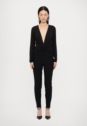 Black long-sleeve jumpsuit with deep V neckline, twisted front detail, slim-fit black pants, and black pointed-toe heels.