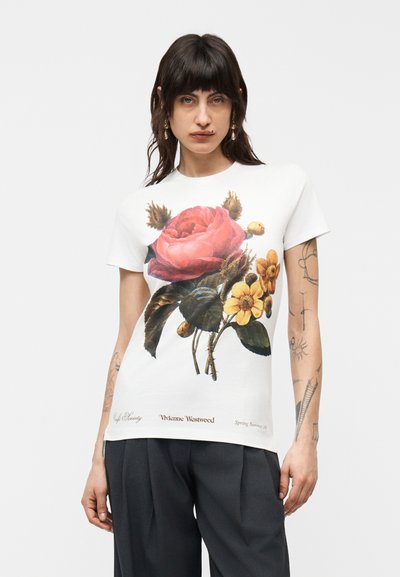 Mujer de cabello oscuro con camiseta blanca con un gran estampado floral en rosa y amarillo y pantalones negros, de pie frente a un fondo liso.