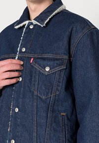 Chaqueta de mezclilla en color azul oscuro, con un cuello de borrego, herrajes plateados y detalles de costura visibles. Bolsillo en el pecho.