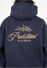 Sudadera de marina con texto bordado en dorado que dice "Prohibido" junto a un diseño de hoja y "10119 BERLÍN" debajo. Tejido suave, corte relajado y capucha amplia.