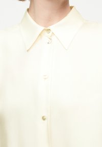 Chemise de couleur crème à col avec quatre boutons fermés, visible du cou à la partie supérieure de la poitrine, portée par une personne aux cheveux courts.