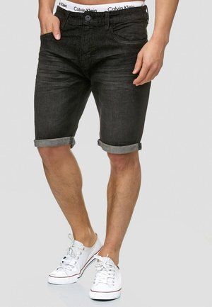 Mężczyzna ubrany w czarne podwinięte krótkie jeansy, białe bieliznę Calvin Klein oraz białe niskie trampki, stojący na szarym tle.