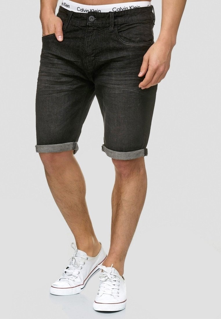 Homme portant un short en jean noir retroussé, un sous-vêtement blanc Calvin Klein et des baskets basses blanches, debout devant un fond gris.
