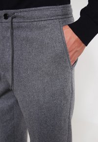 Man wearing gray wool pants with side pocket, hand partially inside pocket, and black long sleeve top visible at wrist.
