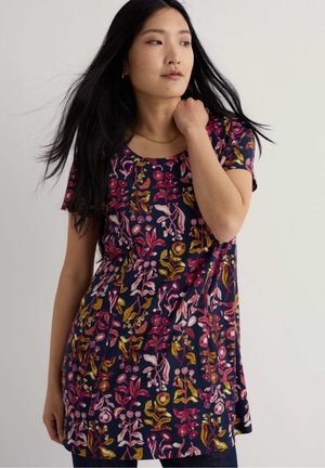 Mladá žena s dlhými čiernymi vlasmi, oblečená v tmavomodrom tričku s krátkym rukávom pokrytom ružovými, červenými a žltými kvetinovými vzormi, pozerajúca sa do strany.