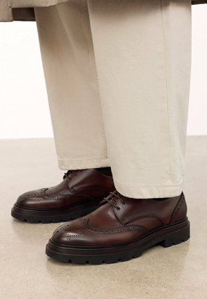 Chaussures brogue en cuir marron avec une semelle texturée à profil bas, des coutures détaillées et des accents perforés. Associées à un pantalon de couleur claire.