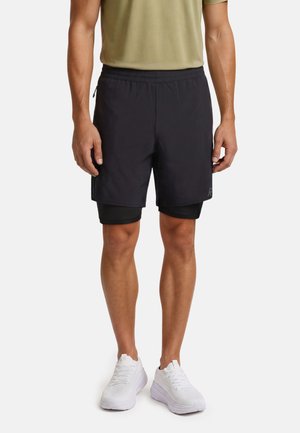 Man draagt zwarte gelaagde sportshorts, olijfgroen shirt en witte hardloopschoenen, staat met armen naast het lichaam.