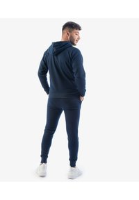 Felpa blu navy con cappuccio e cordino, maniche lunghe e design aderente, abbinata a pantaloni jogger coordinati. Sneakers bianche visibili.
