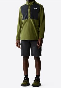 Zielony polarowy sweter z czarnymi ramionami i zapięciem na napy, połączony z czarnymi szortami i butami trekkingowymi, prezentujący sportowy styl.