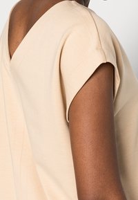 Béžový top s výstřihem do V, krátkými zavinutými rukávy, hladkou texturou a minimálními švy. Boční pohled ukazuje rameno a linii krku.