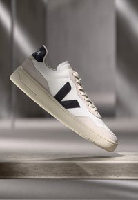 White sneaker with dark blue accents, beige suede details, perforated toe box, and a textured sole. Features a large "V" logo on the side.