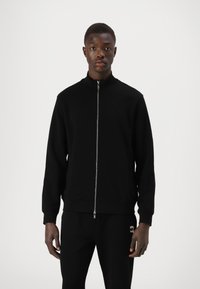 Schwarze Zip-Jacke mit Stehkragen, aus einem weichen Material gefertigt, mit einer dezenten Textur, langen Ärmeln und einer sportlichen Passform.