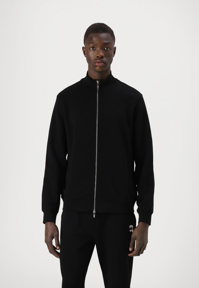 Schwarze Zip-Jacke mit Stehkragen, aus einem weichen Material gefertigt, mit einer dezenten Textur, langen Ärmeln und einer sportlichen Passform.