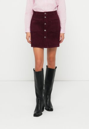 Femme portant une jupe en velours côtelé bordeaux à boutons sur le devant avec poches avant, une chemise rayée rose pâle et des bottes en cuir noires montant jusqu'aux genoux.