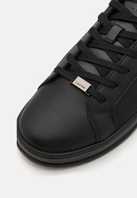 Black leather sneakers with rounded toe, textured side pattern, and black laces. Features a silver "Calvin Klein" logo plate on the side.