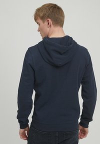 Marineblå hettegenser med kengurulomme, ribbete mansjetter og hette med snor. Stoffet ser mykt og uformelt ut, passende for daglig bruk.