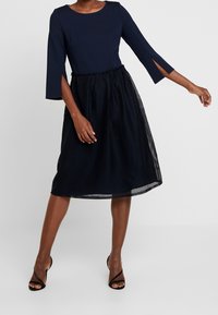 Navy dress with a solid top and a pleated mesh skirt. Features three-quarter sleeves and an elastic waistband for comfort. Black strappy heels.