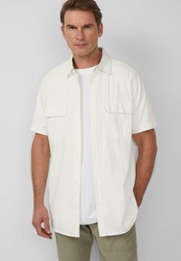 Korte mouwen witte overhemd met een lichte textuur en twee borstzakken, gedragen over een wit T-shirt, gecombineerd met lichtgroene broek.