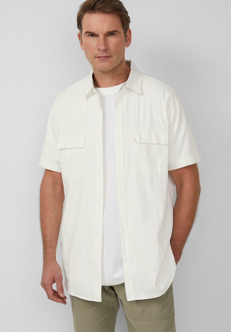 Korte mouwen witte overhemd met een lichte textuur en twee borstzakken, gedragen over een wit T-shirt, gecombineerd met lichtgroene broek.