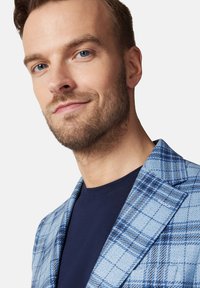 Jasnoniebieska marynarka w kratę, z ciemnoniebieskimi akcentami. Widoczne są wyraźne klapy oraz fakturowany materiał, nałożony na granatową koszulę.