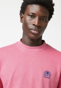 Sudadera de algodón rosa con cuello redondo, que presenta un logo impreso en azul marino de un árbol y rayas en la parte izquierda del pecho.