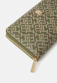 Green patterned wallet with gold zipper, featuring an intricate logo design and textured fabric, rectangular shape, and contrasting stitching.