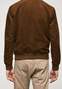 Chaqueta bomber de ante marrón con dobladillo y puños acanalados. La parte trasera presenta un diseño limpio y minimalista sin logotipos ni herrajes visibles.