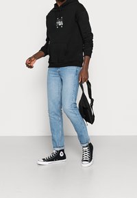 Felpa nera con cappuccio e tasca frontale, jeans azzurri chiari e sneakers nere alte. Porta una piccola borsa a tracolla nera.
