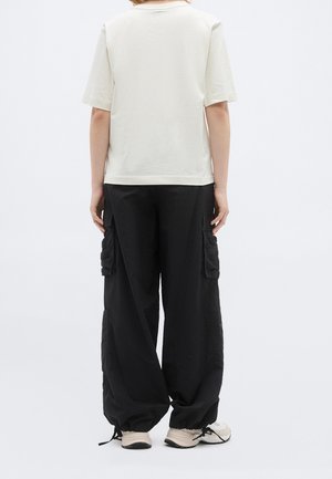 Persona que lleva una camiseta blanca lisa de manga corta y pantalones cargo negros de pierna ancha con dobladillos con cordón, de pie sobre un fondo liso.
