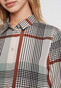 Button-up shirt featuring a grid and striped pattern in black, white, mint green, and rust, with a structured collar and subtle sheen.