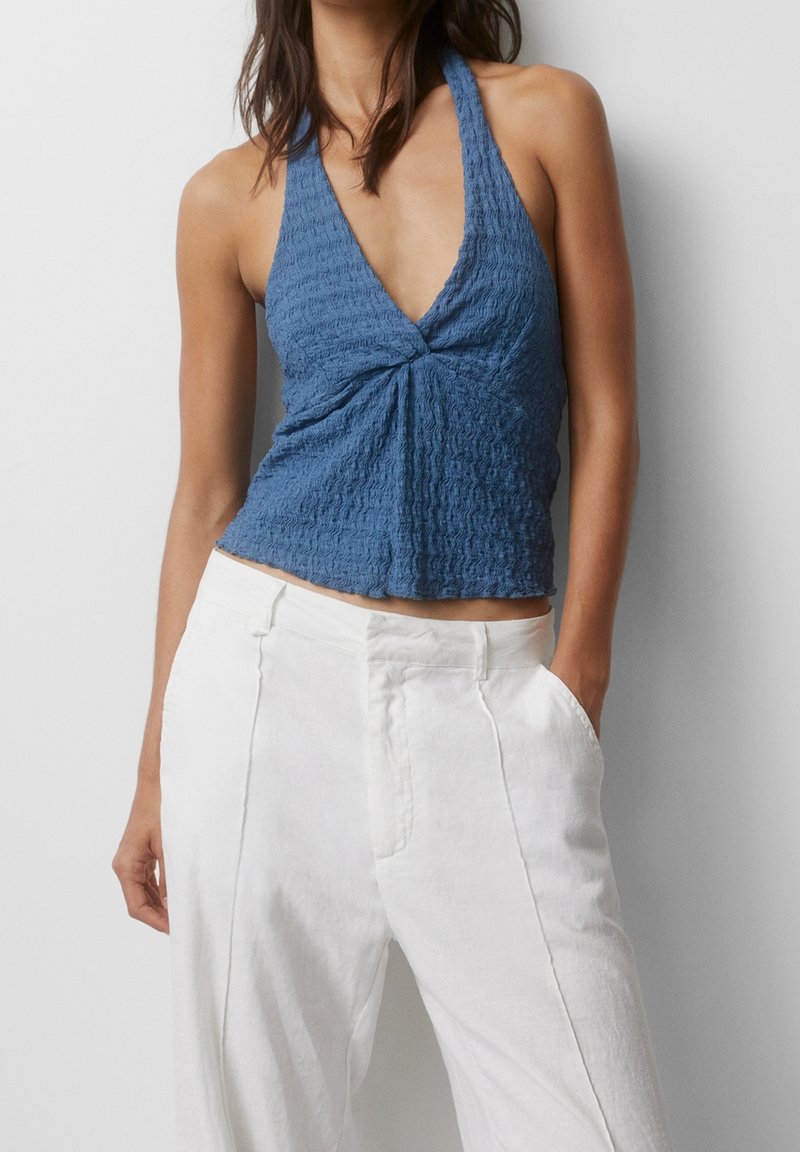 Femme portant un top en tissu bleu texturé à col halter avec un nœud devant et un pantalon blanc taille haute, debout avec une main dans la poche sur fond blanc.