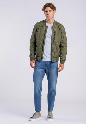 Joven de pie con chaqueta bomber verde oliva, camiseta blanca, vaqueros azules y zapatillas grises sobre un fondo claro y liso.