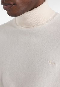 Valkoinen pooloneulepaita, joka on valmistettu pehmeästä neuloksesta. Siinä on ribatumpat kaulus, sileä pinta ja pieni brodeerattu logo rinnassa.