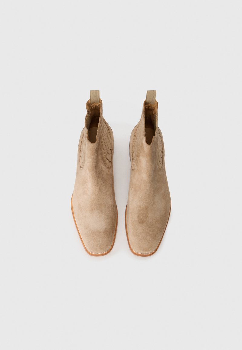 Bottines en daim beige avec orteils pointus, présentant des coutures décoratives et des languettes pour un enfilage facile. Semelle en caoutchouc avec des accents marron clair.
