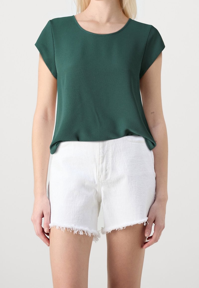 Haut vert à manches courtes avec un col rond, en tissu lisse. Associé à un short blanc à ourlet effiloché.